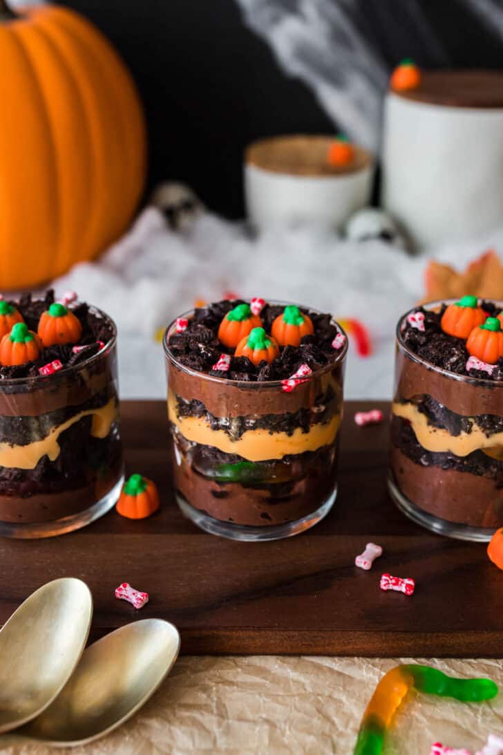 Spooky dessert in glasses made with layers of chocolate and orange colored pudding, crushed cookies and gummy worms. Three cups are arranged on a wooden serving board and topped with candy pumpkins.