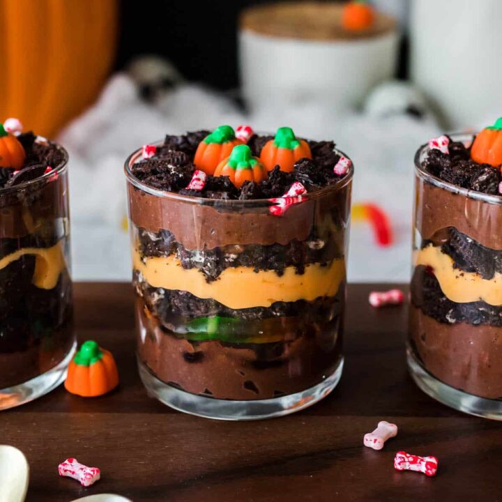 Halloween dirt cups made with layers of chocolate and orange colored pudding, crushed cookies and gummy worms. Three cups are arranged on a wooden cutting board and topped with candy pumpkins.