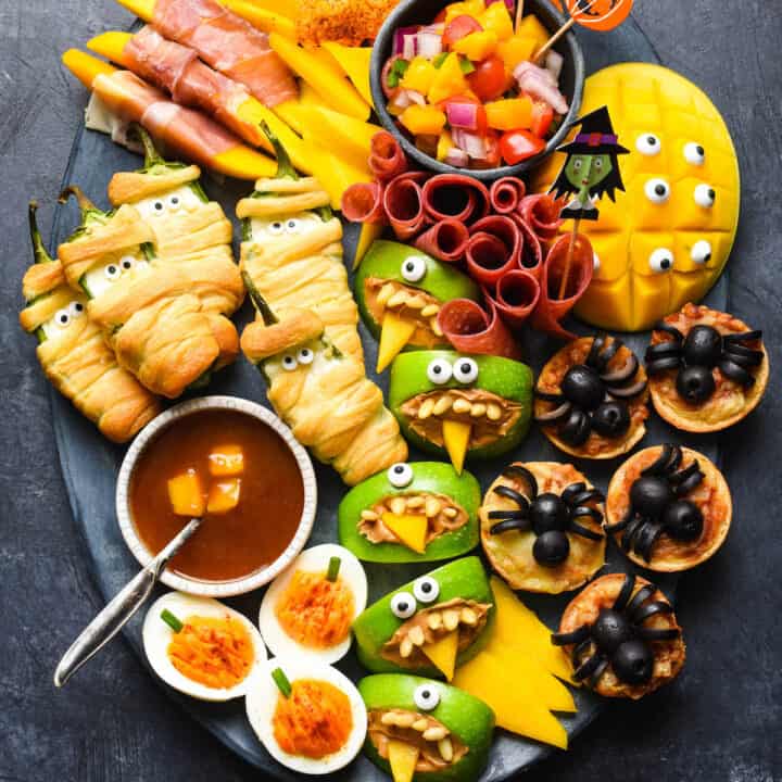 Spooky Halloween food ideas, like jalapeno popper mummies, apple monsters, bat tortilla chips and spider mini pizzas, arranged on a large dark platter on a dark background.