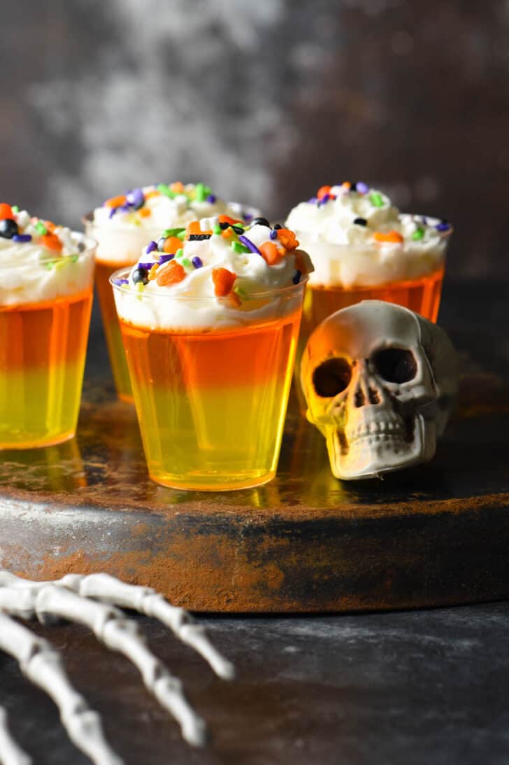 Candy corn gelatin shooters made with layers of yellow and orange gelatin, and whipped cream, topped with sprinkles. Shots are on an upside down tray and the scene is decorated with a fake skull and skeleton hand.