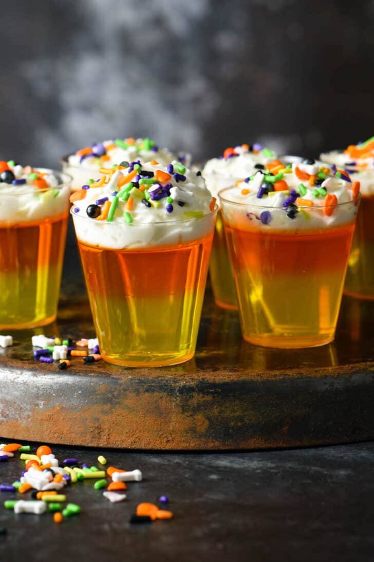 Small plastic shooter glasses filled with layers of yellow and orange gelatin topped with whipped cream and spooky sprinkles.