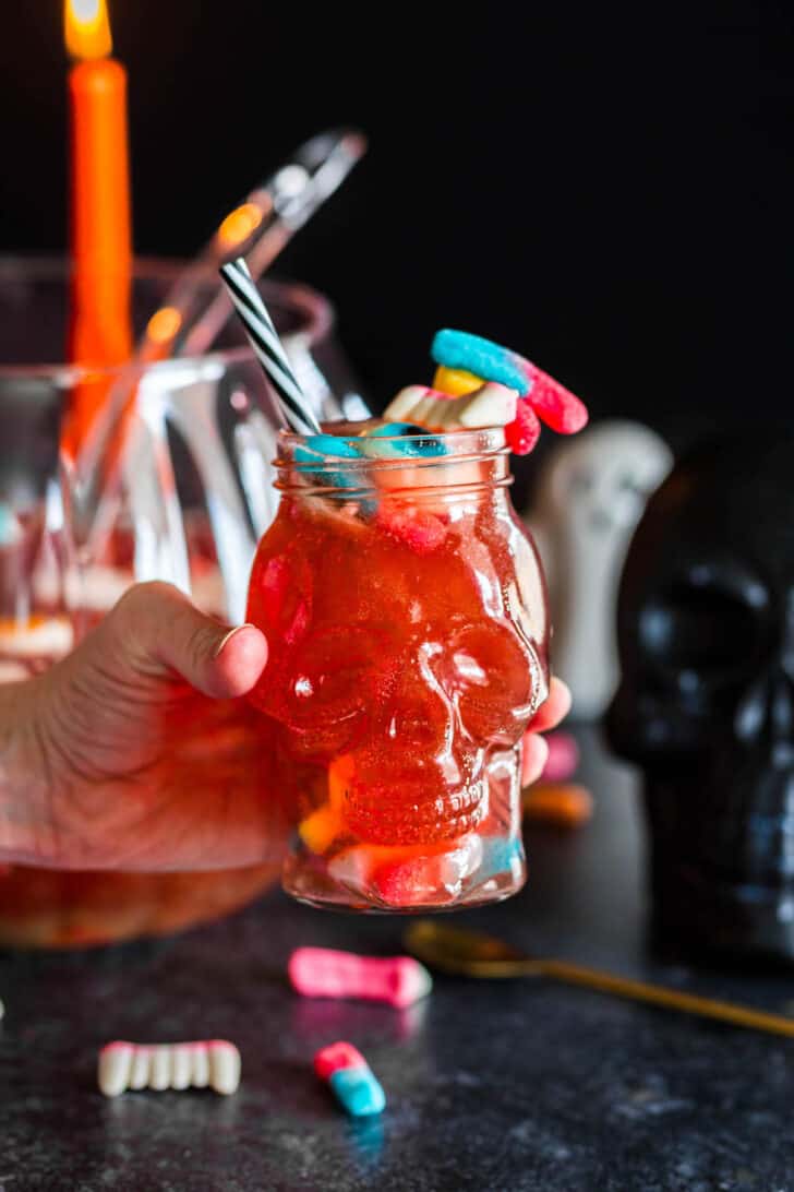 A hand holding a skull shaped tumbler filled with Halloween punch for kids, garnished with candy.