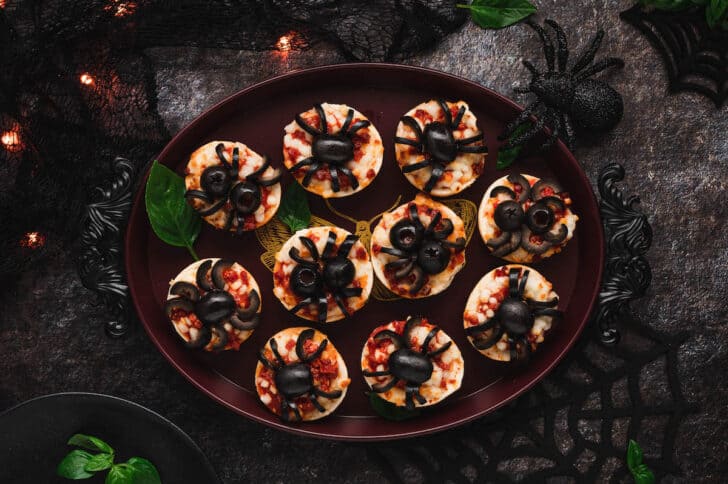 Spider pizzas made with mini pizza bagels and black olives on a maroon tray.