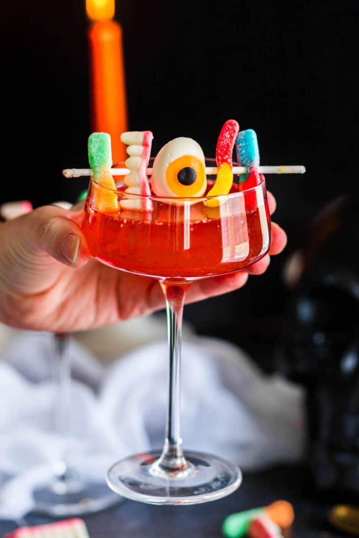 A hand holding a couple glass filled with Halloween party punch, garnished with gummy candies.