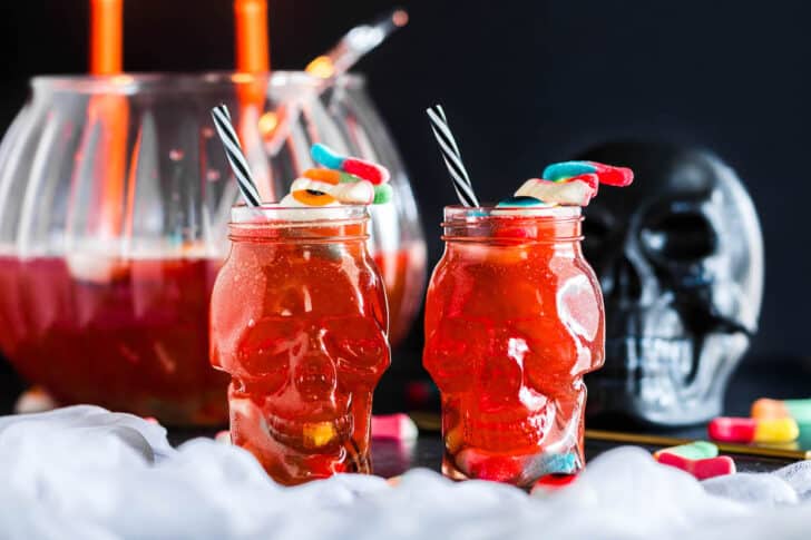 Two skull shaped tumblers filled with Halloween punch for kids, garnished with gummy candies and paper straws.