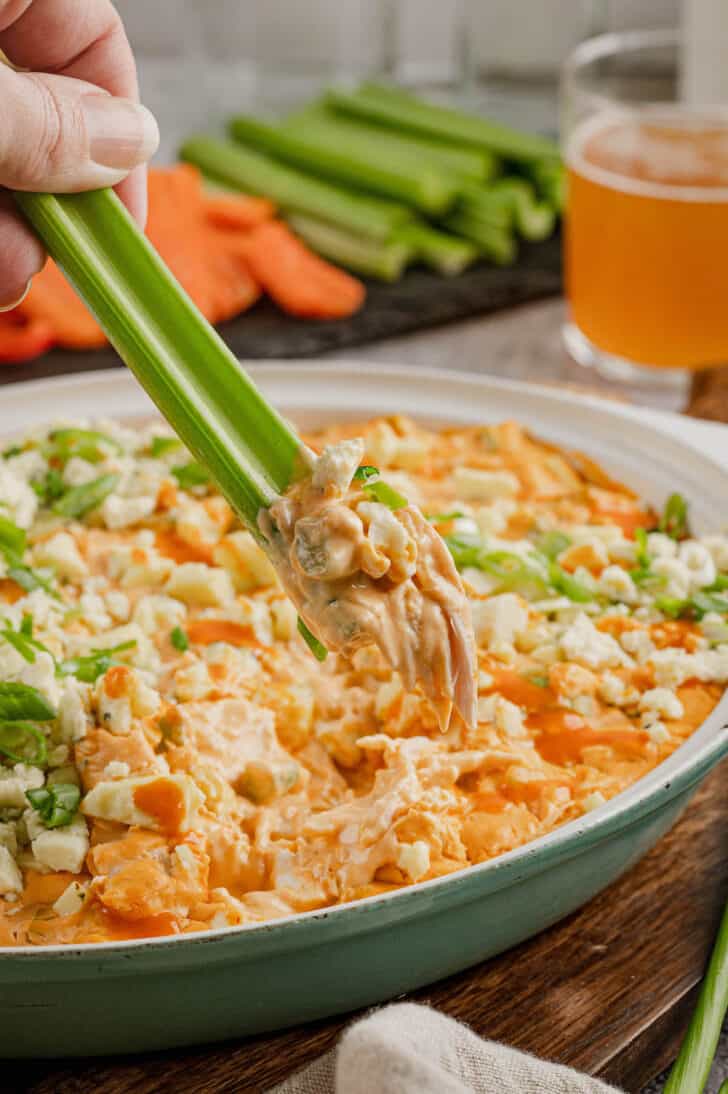A hand dipping a celery stick into a creamy orange chicken and cheese mixture.