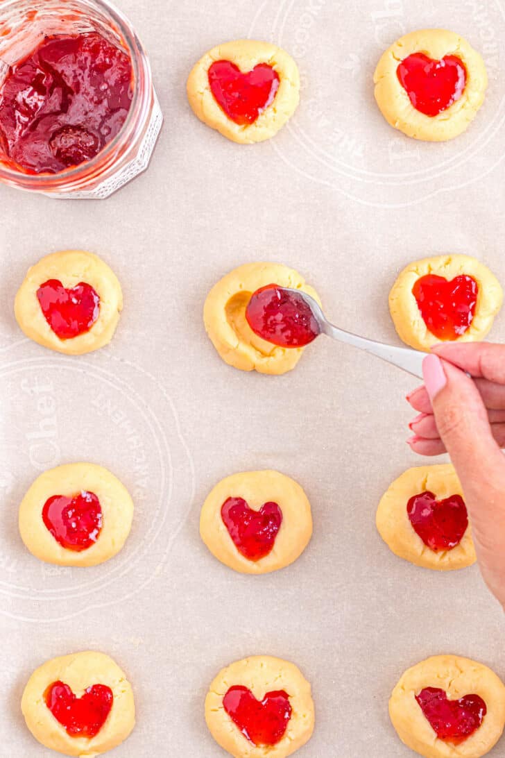 A hand with a small spoon placing jam in strawberry thumbprint cookies.