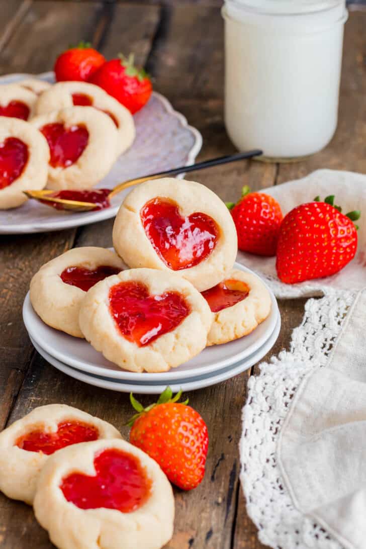 Shortbread rounds filled with strawberry preserves on a stack of small plates on a wooden surface, with fresh strawberries garnishing the scene.