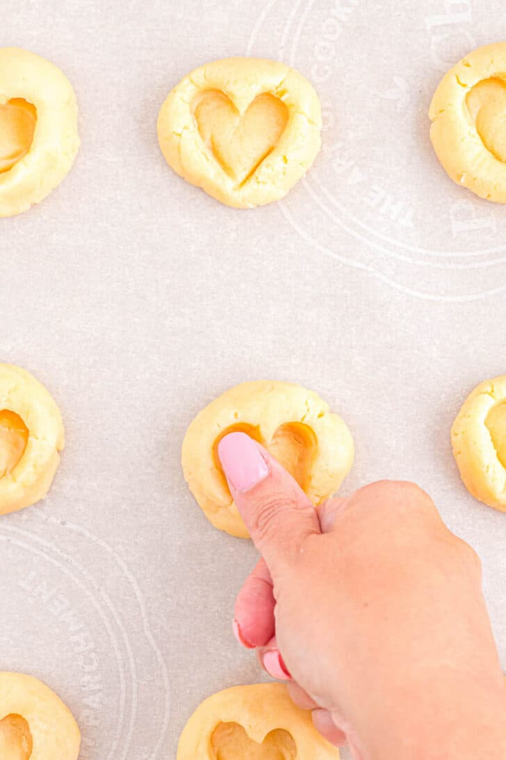 A thumb stamping a heart shape into a shortbread cookie for strawberry jam cookies.