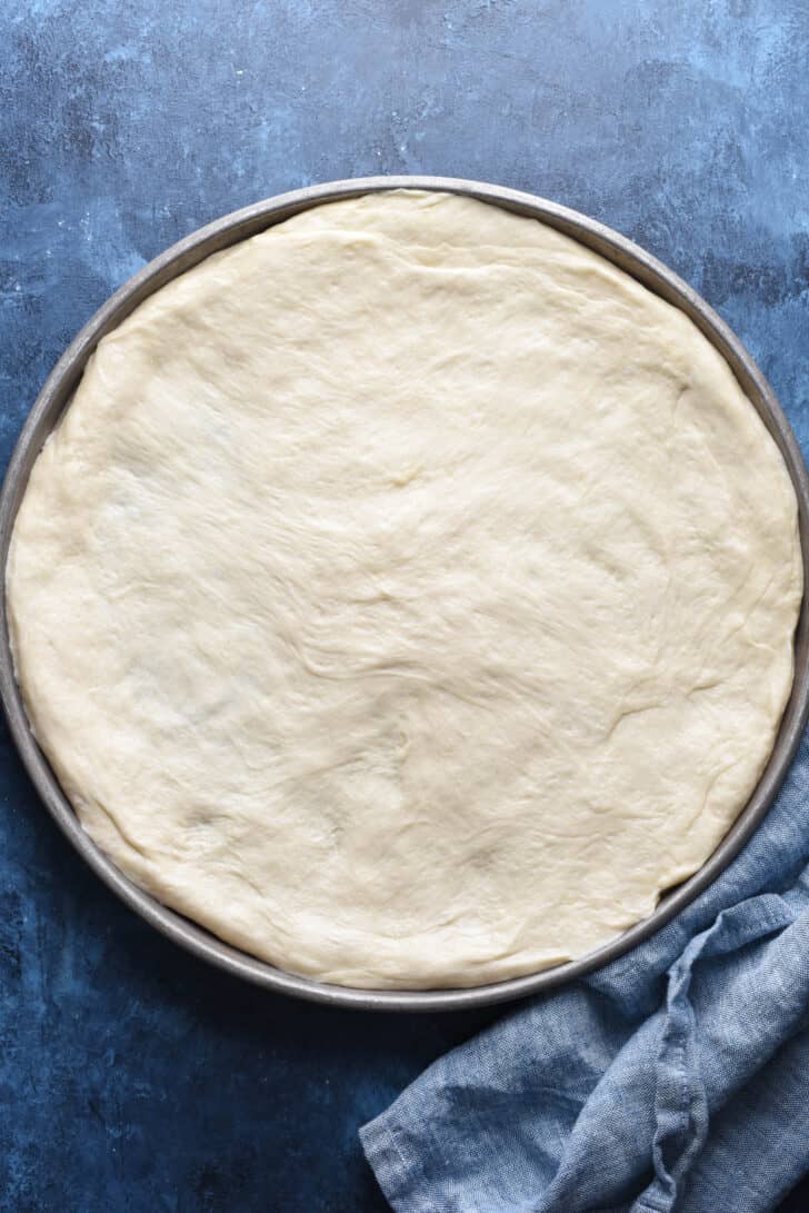 Pizza dough pressed out onto a round pan on a blue background.