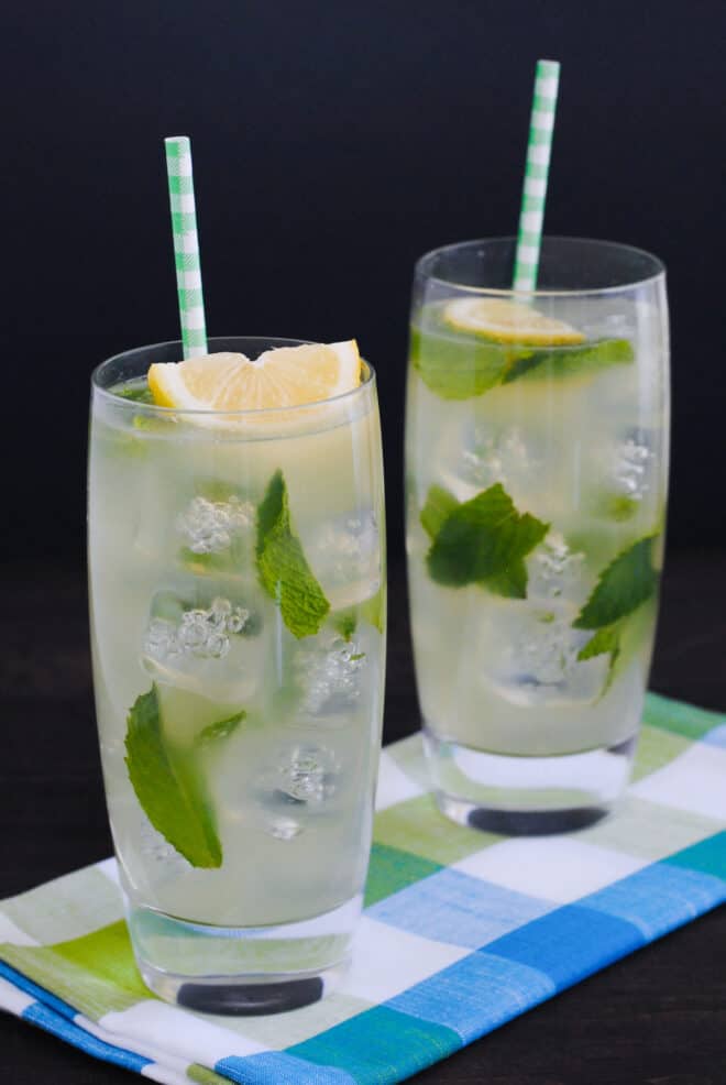 Two tall glasses of mint honey drink over ice, garnished with paper straws and lemon wedges.