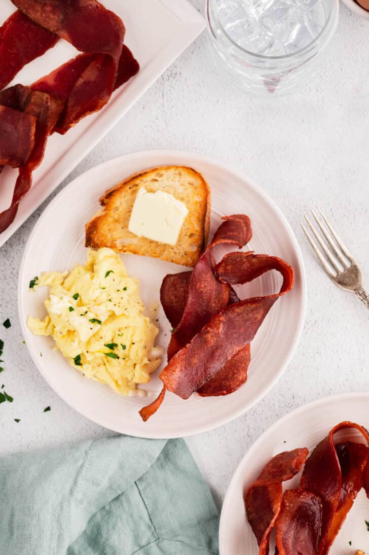 A white breakfast plate with scrambled eggs, toast with butter, and rashers.