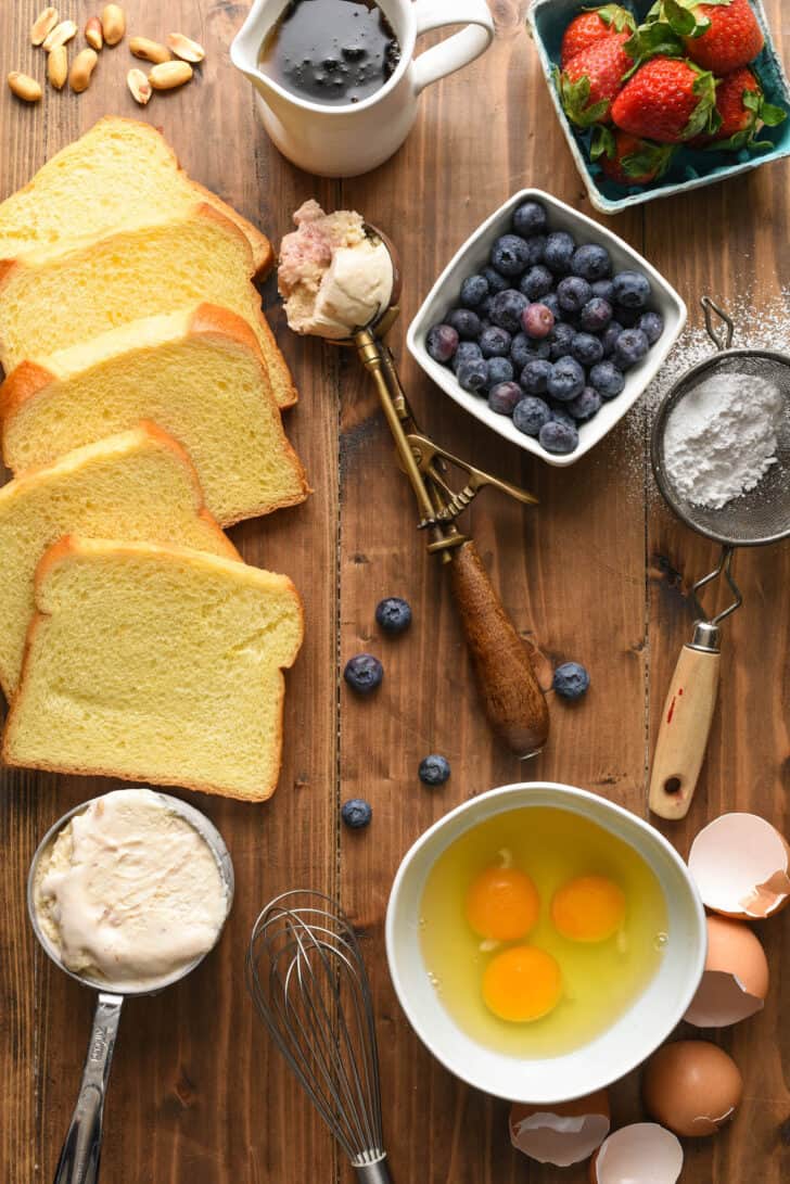 Ingredients laid out on a wooden table, including brioche bread, eggs, dairy, berries and maple syrup.