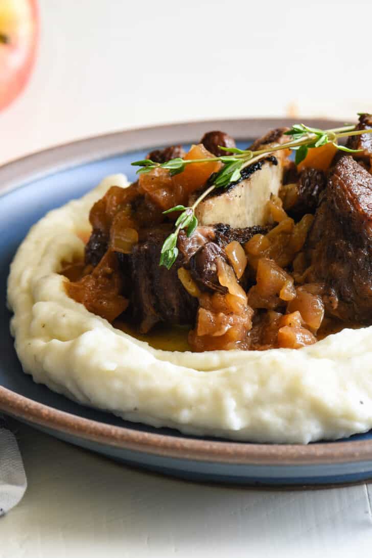 Shallow blue bowl filled with mashed potatoes, topped with braised beef and garnished with thyme.