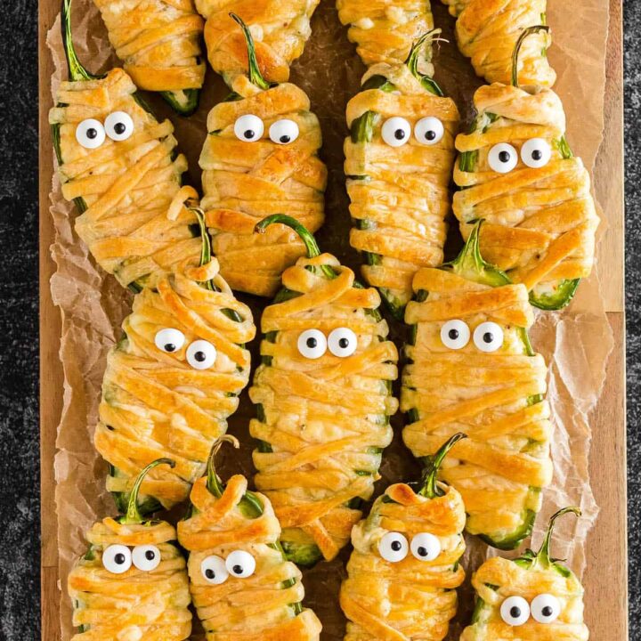 Mummy jalapeno poppers with candy eyeballs on a wooden cutting board.