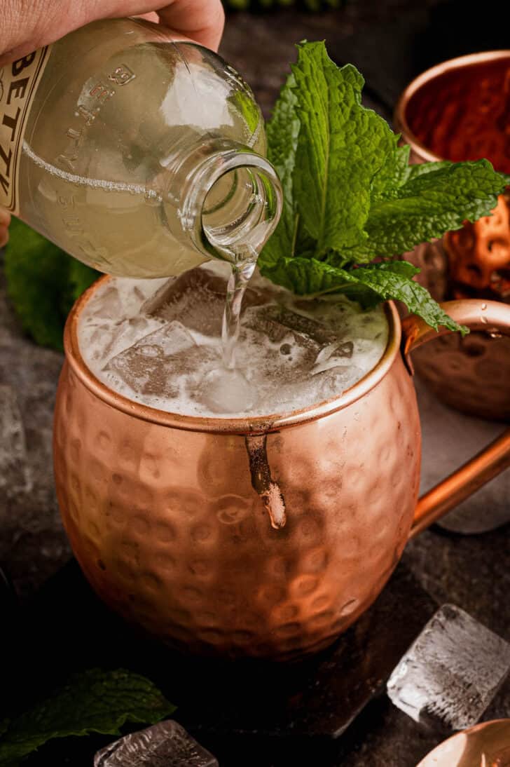 A hammered copper mug filled with a whiskey mule cocktail, garnished with a mint sprig, with a bottle of ginger beer being poured into the mug.