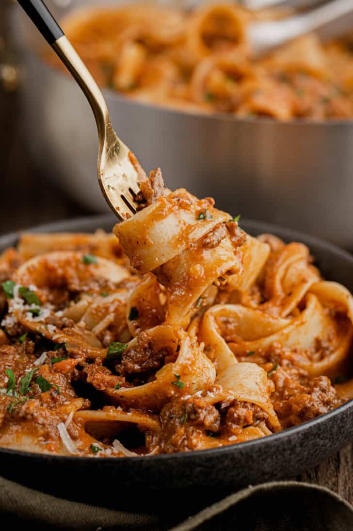 A gold fork twirling pappardelle pasta with meat sauce in a black bowl.