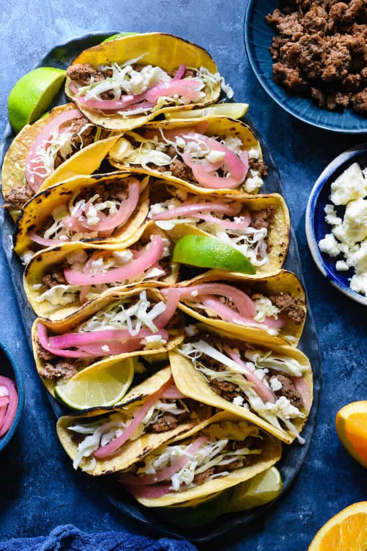 Oval tray filled with lamb tacos garnished with cheese and pickled red onions and lime wedges, on a blue surface.