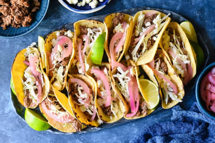 Oval tray filled with ground lamb tacos topped with cabbage, pickled red onions, crumbled white cheese and lime wedges, on a blue surface.
