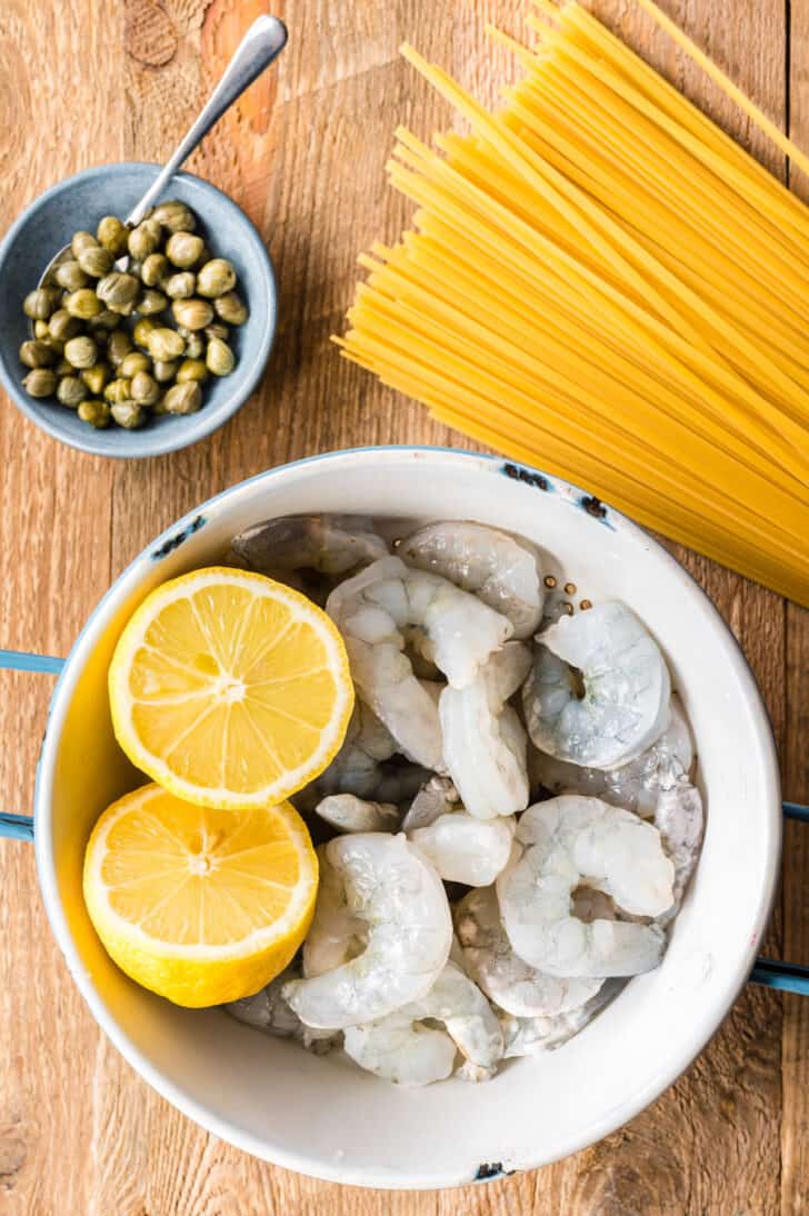 A colander of shrimp and cut lemons, small bowl of capers, and linguine on a wooden surface.