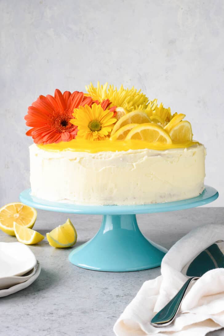 A lemon curd cake decorated with white frosting, lemon slices and yellow and orange flowers, on a teal cake stand.