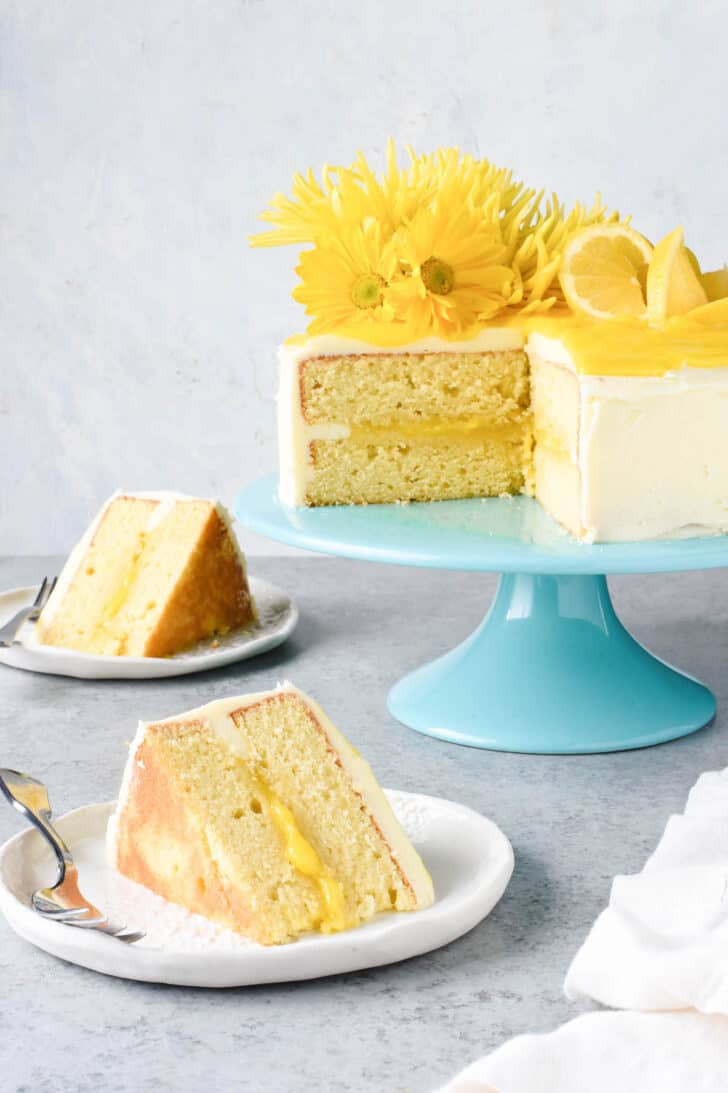 Slices of cake with yellow filling and white frosting on small plates, with the rest of the cake on a teal cake stand.