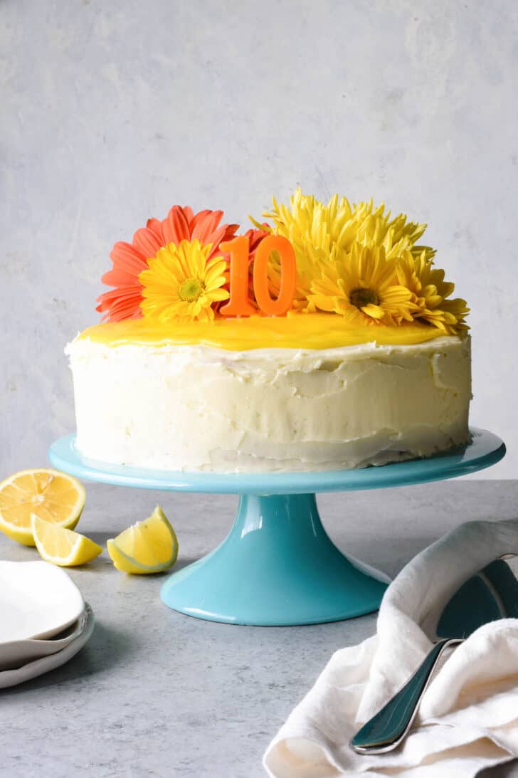 A lemon curd cake decorated with white frosting, orange 1 and 0 candles and orange and yellow flowers, on a teal cake stand.