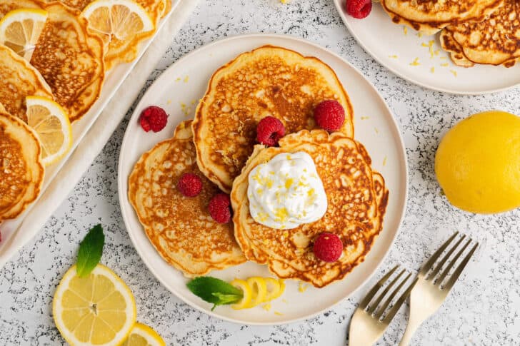 A plate of three pancakes topped with whipped cream, lemon zest and raspberries.