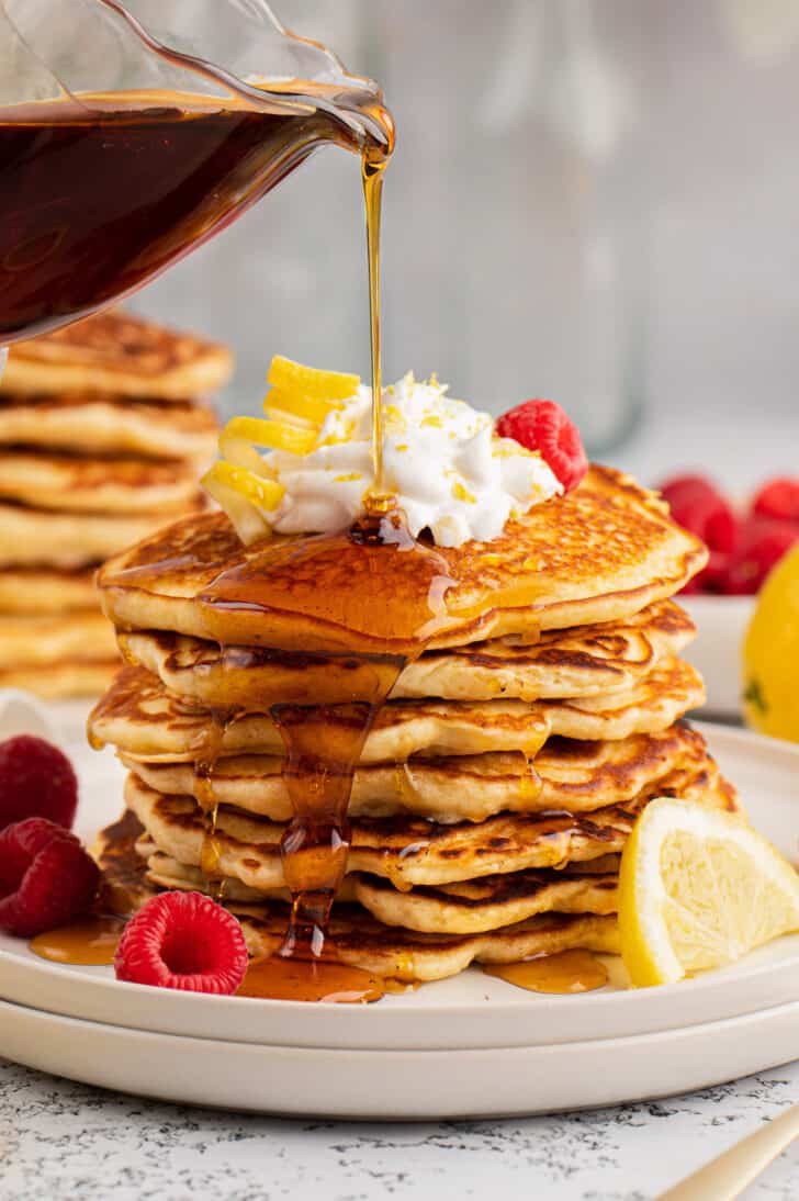 A tall stack of lemon pancakes on a gray plate, garnished with berries and whipped cream, being drizzled with maple syrup.