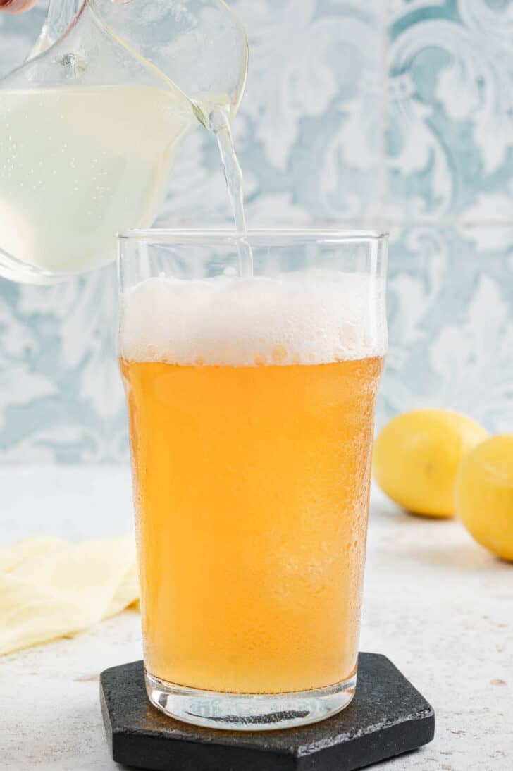 A pint glass of beer being topped off with lemonade from a small pitcher, in front of a patterned blue wall.