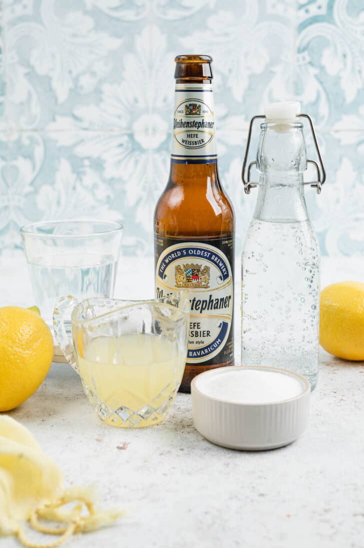Ingredients in front of a patterned blue wall, including a bottle of beer, citrus juice, sugar and simple syrup.