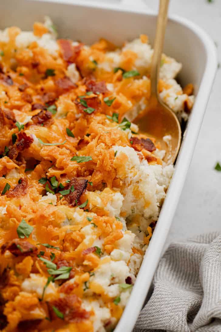 A gold spoon digging into a mashed potato casserole made with cheese and bacon.