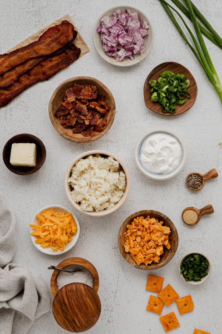 The ingredients needed for a leftover mashed potatoes recipe laid out on a light surface, including bacon, onions, sour cream, butter, cheese, herbs, crackers and mashed potatoes.