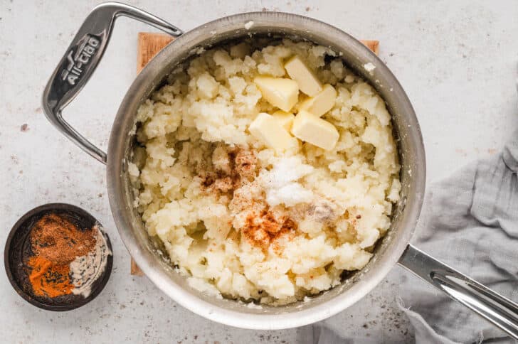 A stainless steel pot filled crushed potatoes, butter and spices.