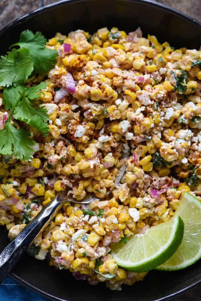 Mexican roasted corn salad in a black bowl, garnished with lime wedges.