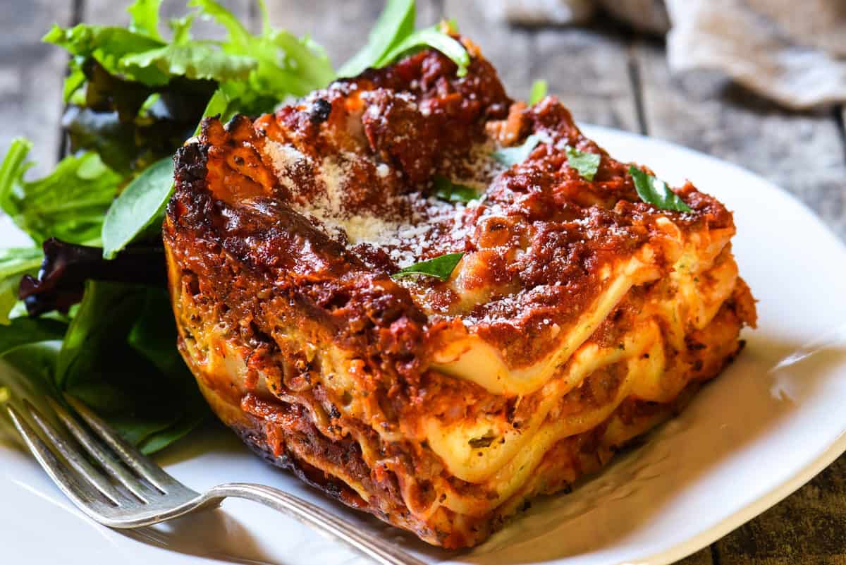 Piece of classic lasagna with cottage cheese with a side salad on a rustic white plate.