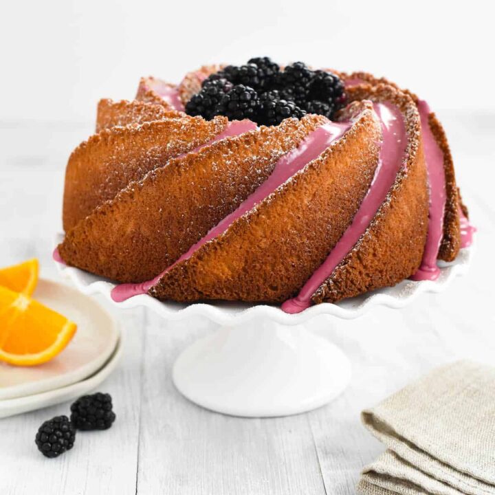 An orange bundt cake decorated with blackberry icing and fresh blackberries, on a white fluted cake stand.