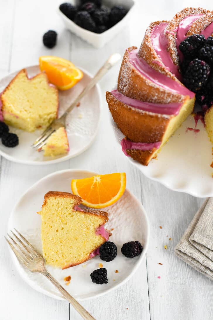 A slice of orange bundt cake on a small white textured plate, garnished with an orange slice and blackberries.