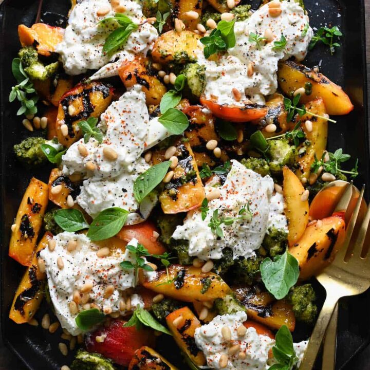 A large black geometric platter piled high with grilled peach burrata salad, garnished with lots of fresh herbs and pine nuts.
