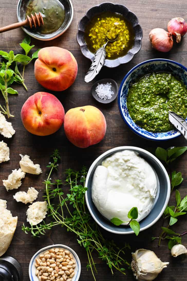 Ingredients laid out on a wooden table, including stone fruit, fresh cheese, pesto, herbs and bread.