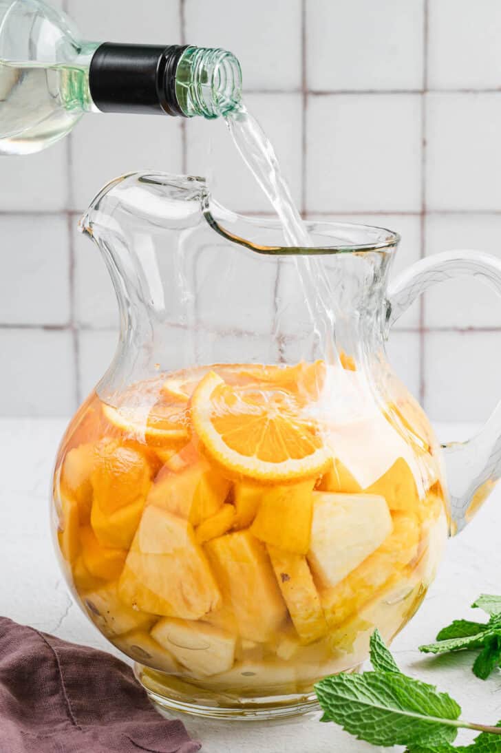A glass pitcher of pineapple mango sangria with white wine being poured into it.