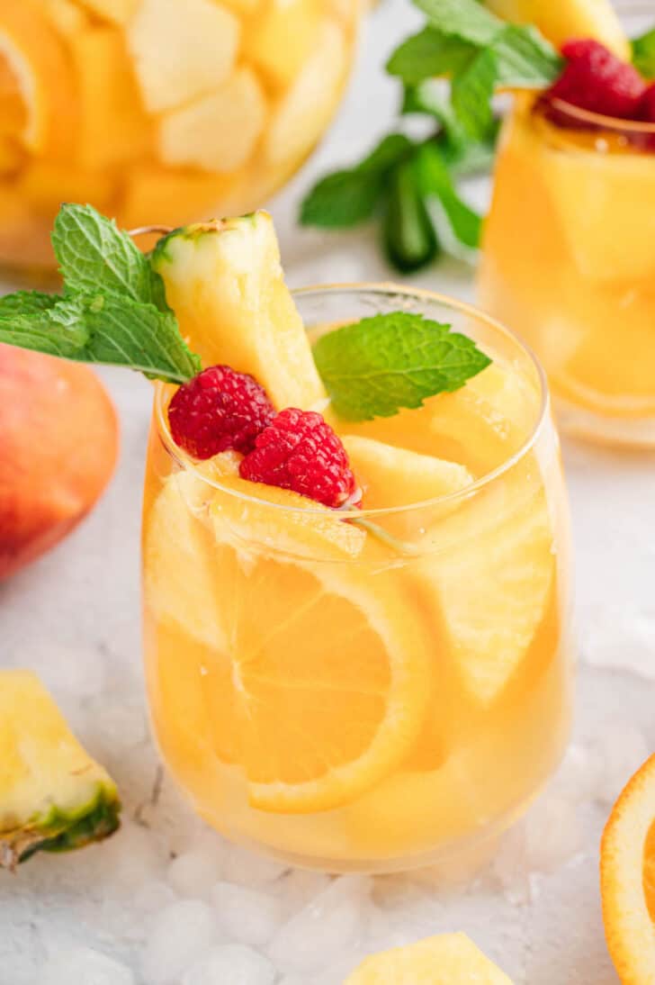 Glasses of wine with citrus fruit and mango over ice, garnished with raspberries and fresh mint sprigs.