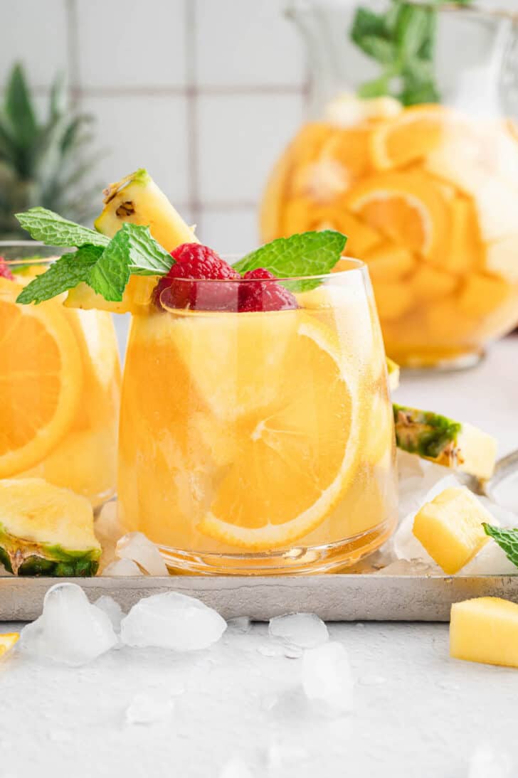 Glasses of pineapple sangria with mango over ice, garnished with raspberries and fresh mint sprigs.