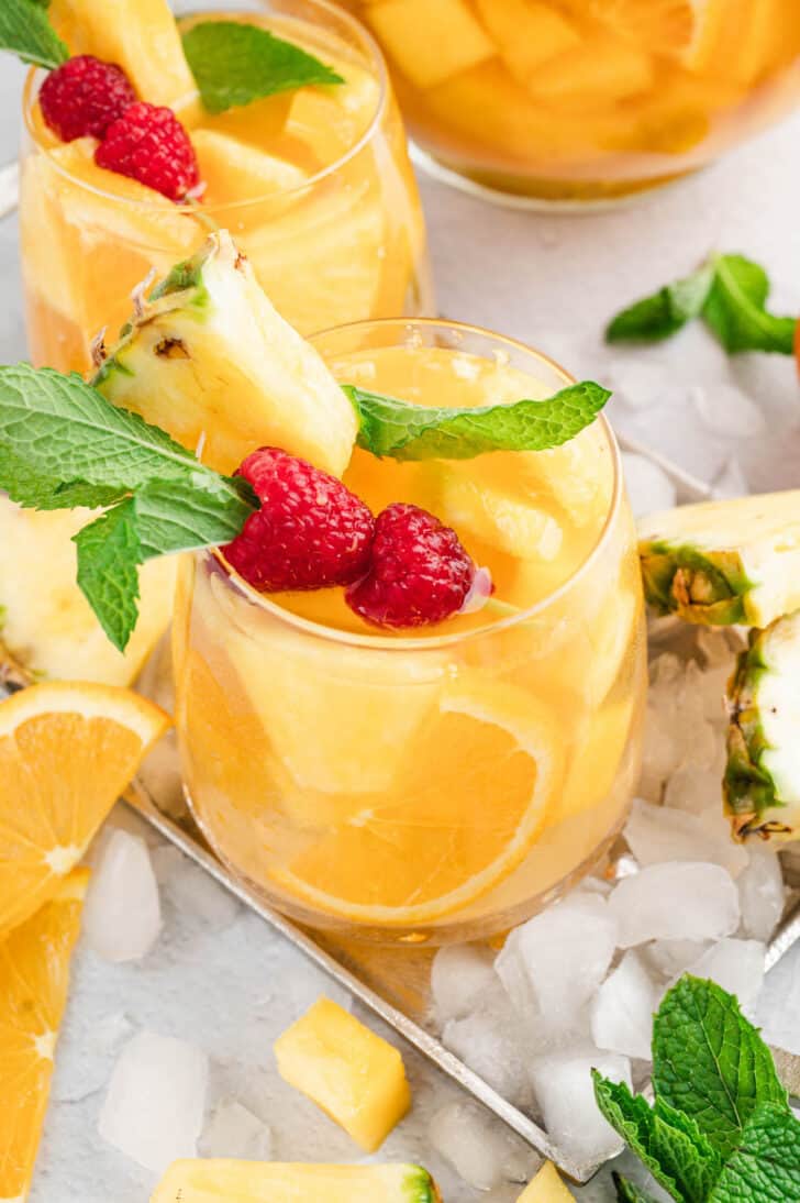 Glasses of pineapple sangria with mango over ice, garnished with raspberries and fresh mint sprigs.