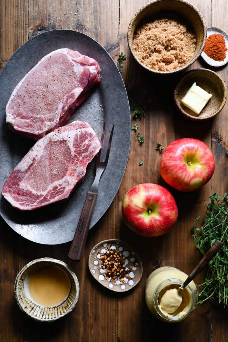Ingredients on a wooden tabletop, including two big pieces of meat, apples, spices, thyme, mustard, brown sugar and butter.