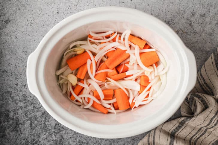 A white slow cooker insert with sliced carrots, onions and garlic inside.