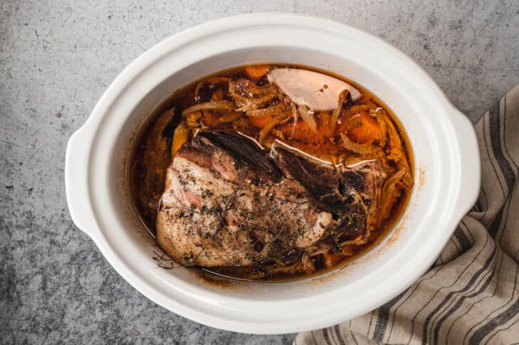 A white slow cooker insert with sliced carrots, onions and garlic with a pork roast on top inside, after the cook.
