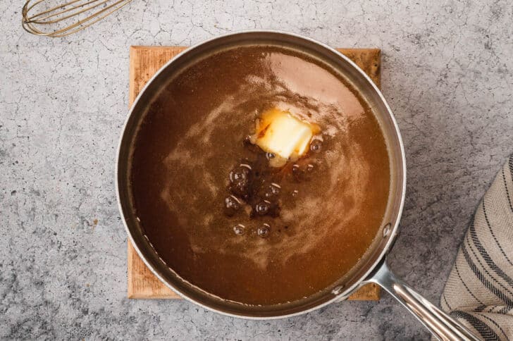 A stainless steel saucepot filled with brown gravy, with a pat of butter and a splash of soy sauce added to it.