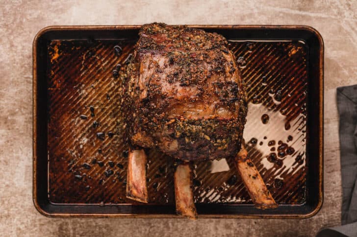 A cooked standing rib roast with a dry rub for prime rib on it, on a rimmed baking pan.