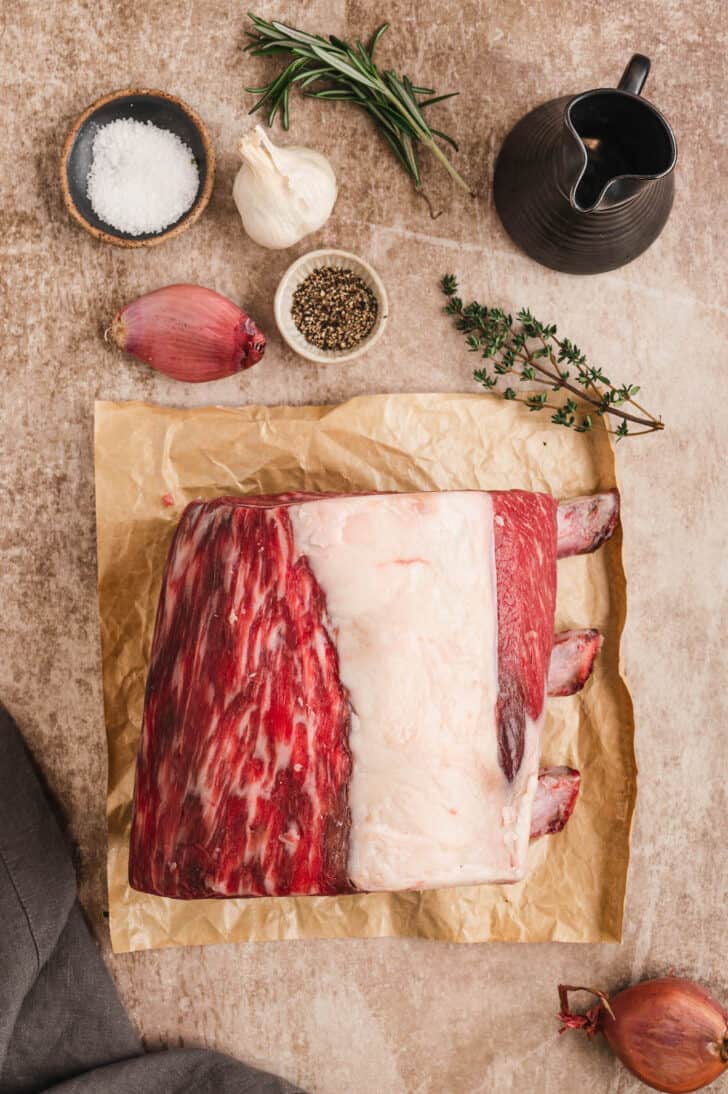 Ingredients on a light brown background including a large piece of bone-in red meat, thyme, rosemary, garlic, shallot and salt and pepper.